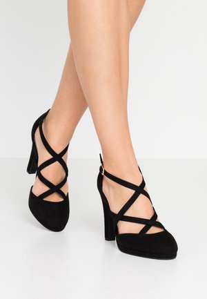 Beine mit schwarzen High Heels mit mehreren überkreuzten Riemen und einer Schnalle vor hellem Hintergrund.