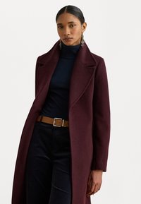 Lungo cappotto bordeaux con colletto a risvolto, realizzato in tessuto testurizzato. Indossato sopra un dolcevita blu navy, abbinato a una cintura beige e pantaloni scuri.