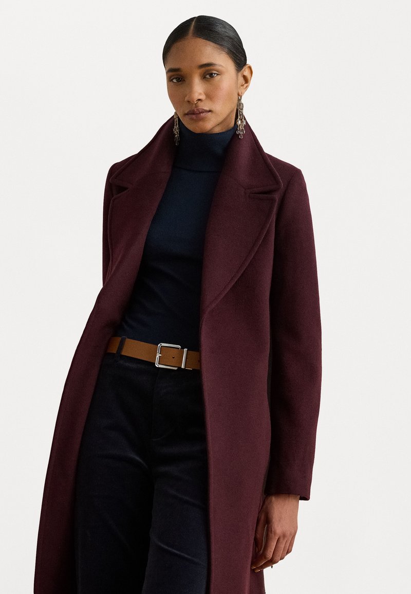 Lungo cappotto bordeaux con colletto a risvolto, realizzato in tessuto testurizzato. Indossato sopra un dolcevita blu navy, abbinato a una cintura beige e pantaloni scuri.