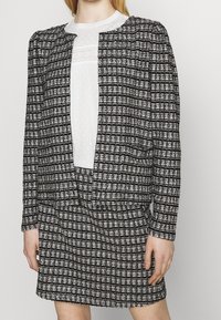 Černo-bílý kostkovaný blazer přes bílou blůzu s texturovanými detaily, obsahující nabírané rukávy a odpovídající kostkovanou sukni.