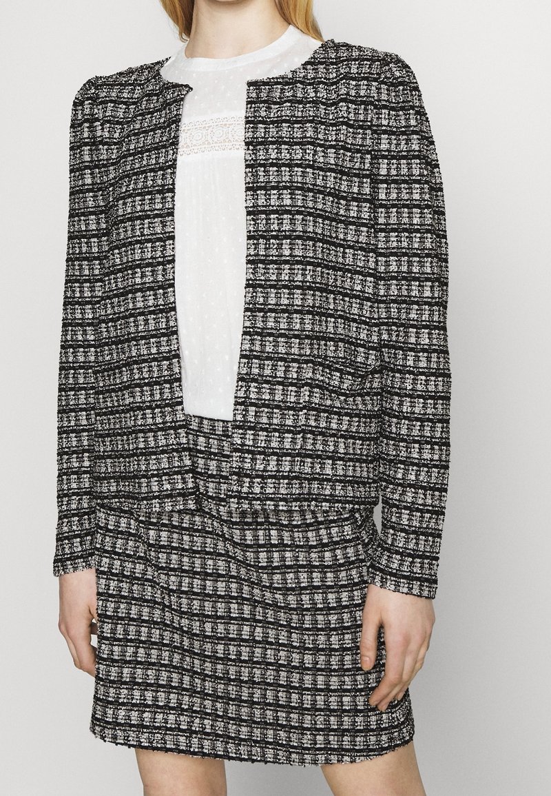 Černo-bílý kostkovaný blazer přes bílou blůzu s texturovanými detaily, obsahující nabírané rukávy a odpovídající kostkovanou sukni.