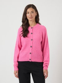 Mulher com cabelo longo e escuro, vestindo um cardigan rosa-choque com botões e calças pretas, de pé contra um fundo claro e liso.