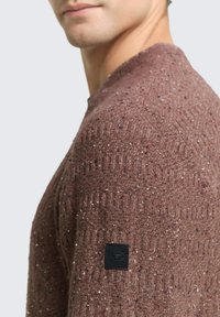 Pull marron texturé avec un design à côtes, des éclats de rouge et de blanc, et une petite étiquette logo noire sur l'épaule gauche.