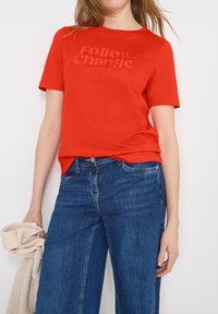Femme portant un t-shirt rouge avec le texte « Follow Change Dance », un jean bleu, tenant un pull en tricot beige sur fond blanc.