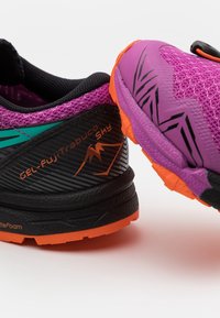 Zapato con parte superior de malla negra y morada con acentos en color aqua, suela naranja, superficie texturizada y la marca "GEL-FujiTrabuco Sky".