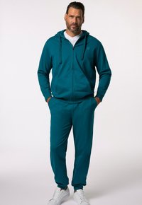 Tealfärgad zip-hoodie med justerbara snoddar, tillsammans med matchande joggers. Tillverkad av mjukt material, har sidofickor och vita sneakers.