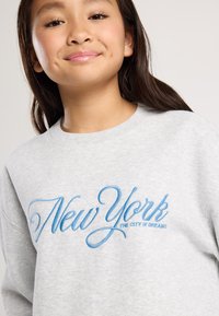 Nuori henkilö, jolla on pitkät tummat hiukset, käyttää vaaleanharmaata hupparia, johon on kirjailtu sinisellä tekstillä "New York Unelmien Kaupunki."