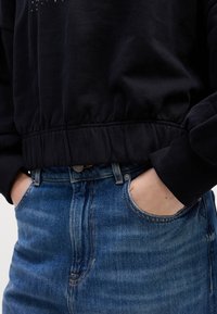 Personne portant un jean en denim bleu taille haute, les mains dans les poches et un sweat court noir avec une taille élastique.