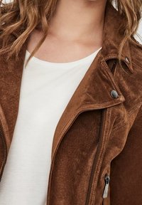 Hnědá semišová bunda se zipem v přední části, stříbrnými akcenty a s vykrojeným límcem, která je navrstvená přes bílou košili.