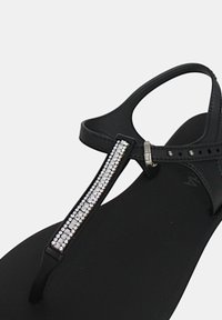 Sandalo infradito nero con suola piatta, una fila decorativa di strass lungo la cinghia e una fibbia regolabile alla caviglia.