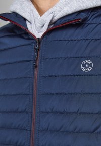 Marineblå pufferjakke med quiltet design, grå hættetrøje under, kontrasterende lynlås og cirkulært logo på brystet. Glat overflade, afslappet pasform.