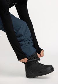 Black snow boot with rubber sole and lacing system, worn with dark blue ski pants and black long-sleeve top, being tightened.