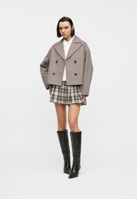 Γυναίκα που φοράει γκριζοπράσινο κοντό διπλόψηλο σακάκι, λευκό πουκάμισο, καρό μίνι φούστα και μαύρα μπότες μέχρι το γόνατο, στέκεται μπροστά σε λευκό φόντο.