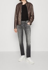 Hombre con chaqueta de cuero marrón, camisa negra, jeans gris descolorido y zapatillas blancas de pie contra un fondo liso.