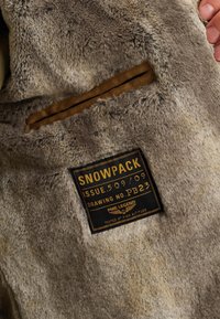 Hnedá kožušinová bunda s béžovou podšívkou a obdĺžnikovou čiernou etikettou so zlatým textom, ktorý uvádza "SNOWPACK" a pokyny na údržbu.