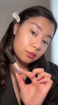 NARS concealer-tub, transparent med svart lock, hålls i en hand. Modellen har en jämn, naturlig hudstruktur och ljust brunt hår med en hårklämma.