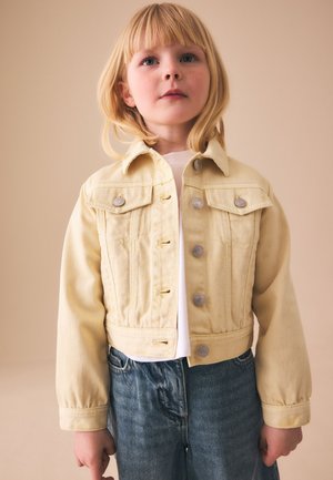 Unge barn med blondt hår iført en lys gul jakke og blå jeans, stående mod en beige baggrund.