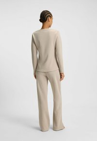 Vrouw met gevlochten haar, gekleed in een beige, textuurrijke lange mouwtop en bijpassende wijde broek, die blootsvoets met de rug naar de camera staat op een witte achtergrond.