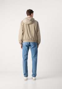 Lys beige hættejakke med elastiske manchetter og kant, sammen med lyseblå lige ben jeans og hvide sneakers, set fra ryggen.