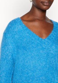 Persona con pelle scura che indossa grandi orecchini a cerchio d'oro e un maglione a V di un vivace blu, mostrata dalle spalle alle labbra.