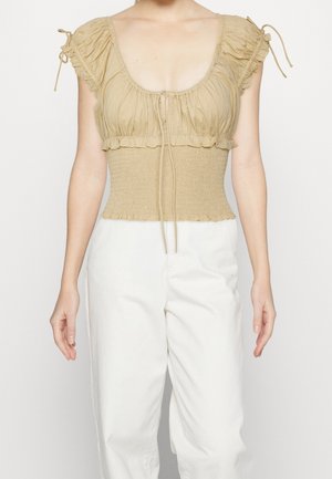 Femme portant un crop-top beige froncé avec manches bouffantes et un pantalon blanc taille haute sur un fond uni.