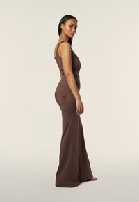 Abito marrone aderente con pantaloni larghi, design senza maniche, tasche laterali e una texture liscia, indossato da una modella su uno sfondo neutro.