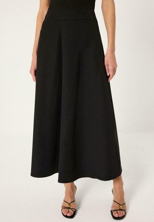 Femme portant une jupe midi taille haute noire en ligne A avec des talons noirs ouverts à brides, debout devant un fond uni.