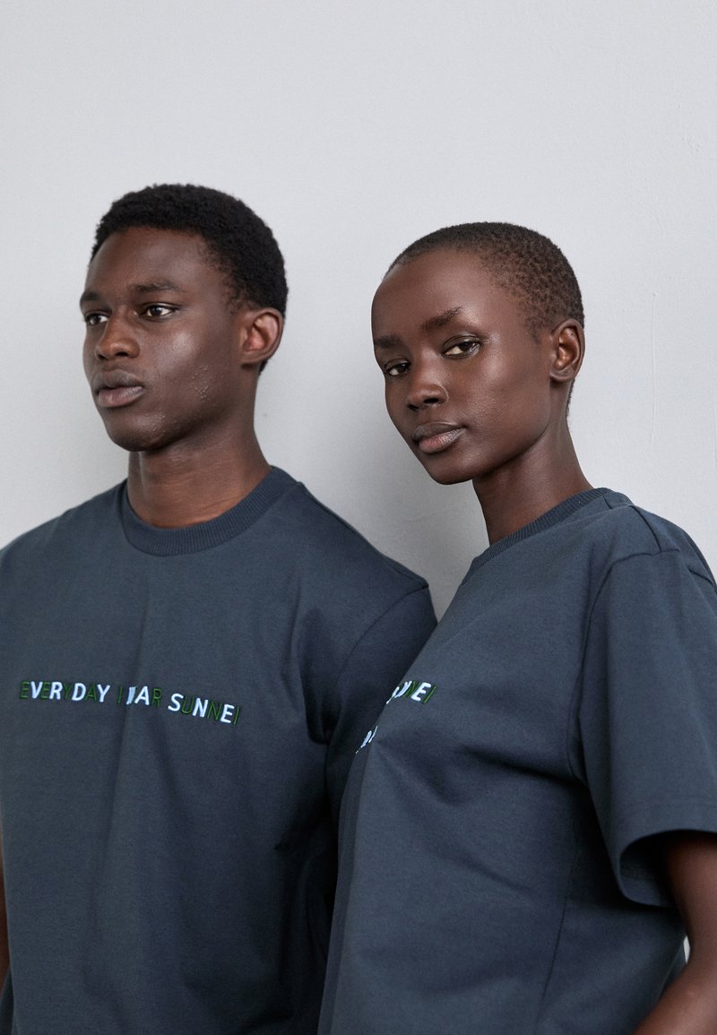 Two individuals wear dark gray cotton t-shirts featuring the text "EVERYDAY SUNNE" in contrasting green letters across the chest.