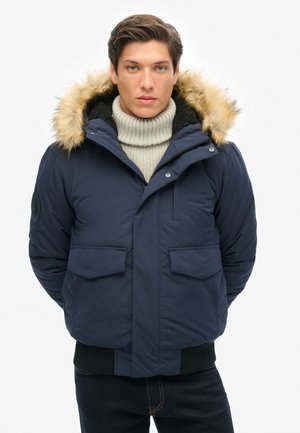 Chaqueta aislante azul marino con capucha forrada de piel, grandes bolsillos frontales y un dobladillo de punto negro. Cuenta con cierre de cremallera y botones a presión.