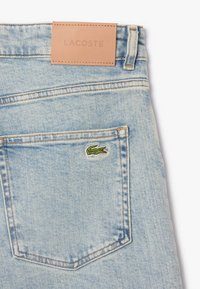 Jeans in denim azzurro chiaro con un'etichetta in pelle beige che riporta la scritta "LACOSTE" in vita e il logo del coccodrillo ricamato di colore verde sulla tasca posteriore.