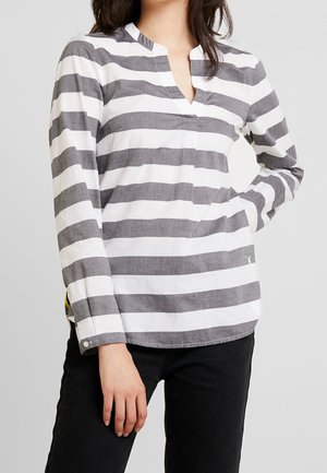Vrouw draagt een lange mouwen, grijs-wit horizontaal gestreepte V-hals blouse met zwarte broek, poseert met één hand op de heup.