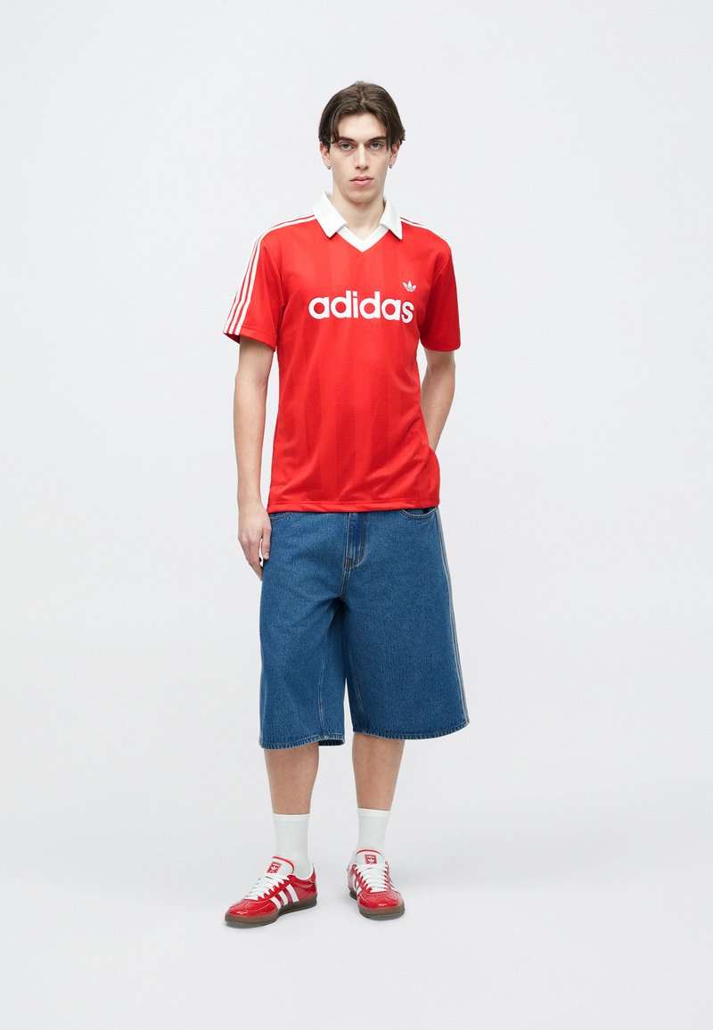 Jeune homme debout portant un t-shirt rouge Adidas, un short long en denim, des chaussettes blanches et des baskets rouges Adidas, sur un fond uni.
