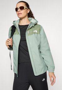 Jachetă impermeabilă verde deschis, cu glugă, având accente verzi închise și un logo. Asortată cu un top negru și pantaloni ajustați.