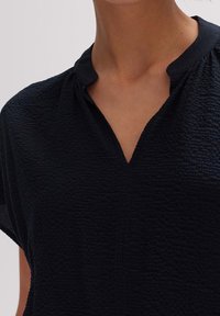 Close-up van een persoon die een gestructureerde zwarte V-hals top met een kleine staande kraag en korte mouwen draagt.