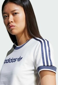 Bílé tričko s krátkým rukávem, které má na ramenou námořnické modré tři pruhy, kulatý výstřih a na přední straně potisk loga Adidas.