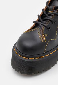 Zapato de cuero negro con costuras amarillas, punta redondeada y suela gruesa de goma negra con un patrón de relieve pronunciado. Diseño con cordones.