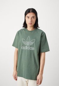 Zielona bawełniana koszulka z białym logo Adidas i wzorem trzech liści z przodu. Krótkie rękawy i luźny krój.