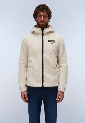Mann mit lockigen Haaren und Bart, trägt eine beige Fleecejacke mit Kapuze und dunkelblaue Jeans, steht vor einem schlichten Hintergrund.