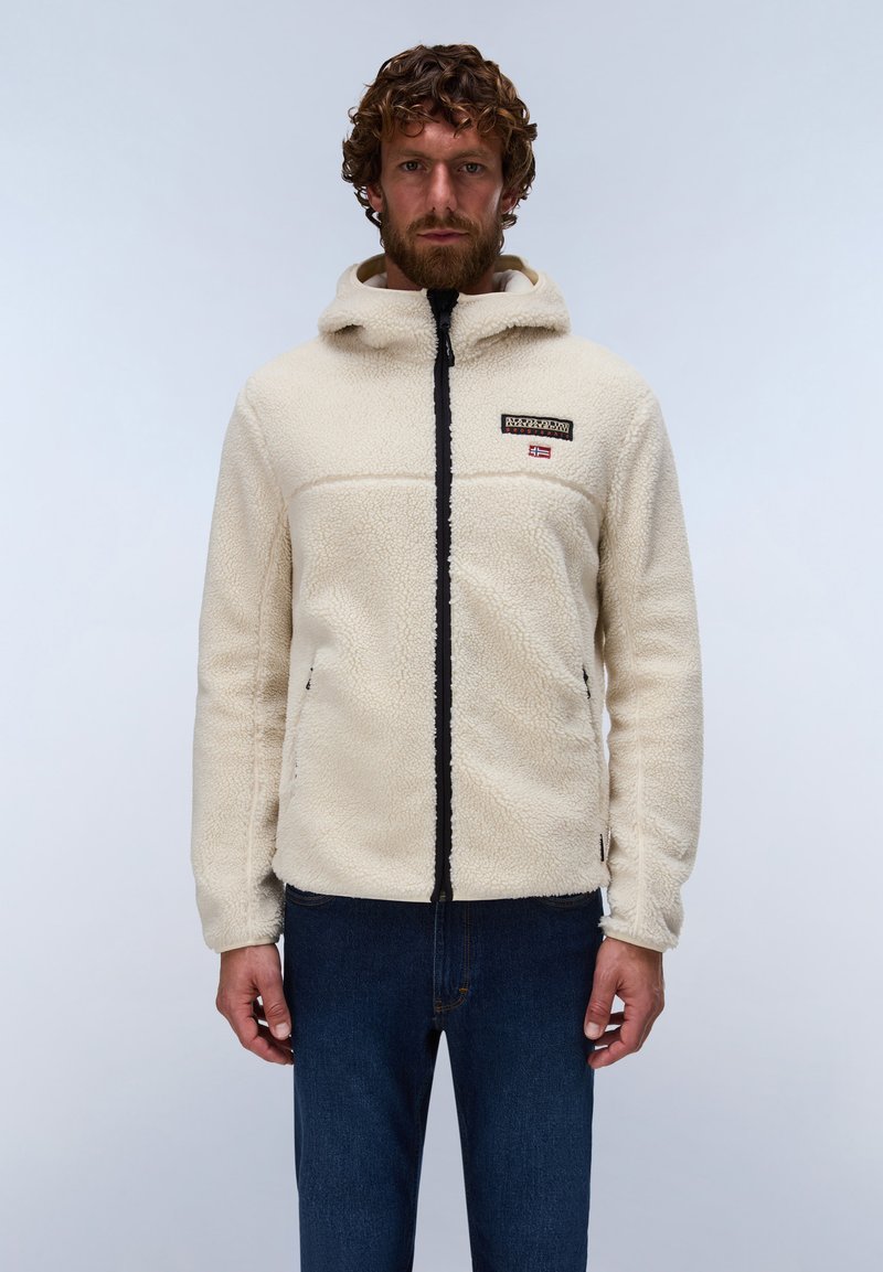 Mann mit lockigen Haaren und Bart, trägt eine beige Fleecejacke mit Kapuze und dunkelblaue Jeans, steht vor einem schlichten Hintergrund.
