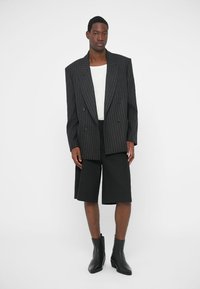 Dwurzędowa marynarka w paski w kolorze czarnym i szarym, zestawiona z dopasowanymi czarnymi szortami i eleganckimi czarnymi botkami za kostkę. Gładki materiał, wyraźne linie.