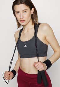 Sutia desportivo cinzento com decote redondo, apresentando o logótipo da Adidas. Corda de saltar preta segurada com ambas as mãos, acompanhada de pulseiras pretas. Leggings bordeaux.