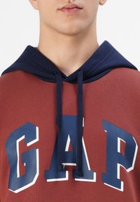 Viininpunainen huppari, jossa on laivastonsininen huppu ja kiristysnyörit. Rintakehässä suuri valkoinen ja sininen "GAP"-logo. Puuvillamateriaalia, sileästä kankaasta.