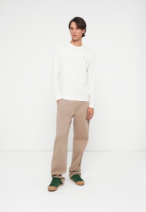 Noor mees seisab, käed beežide pükste taskutes, seljas valge pikakäiguline särk ja rohelised tennisetossud, neutraalne näoilme, ühtlane valge taust.