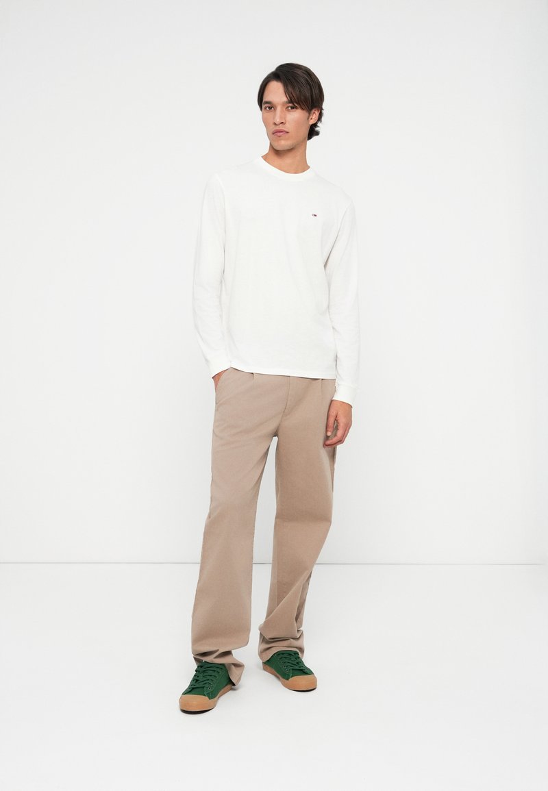 Ung mand står med hænderne i lommerne på beige bukser, iført en hvid langærmet skjorte og grønne sneakers, neutral ansigtsudtryk, ensfarvet hvid baggrund.