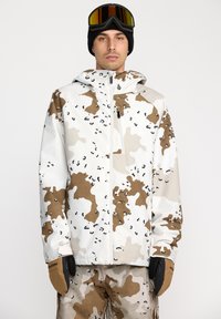 Camouflagejas in wit, bruin en beige met een ritssluiting aan de voorkant, verstelbare capuchon en elastische manchetten. Gecombineerd met bruine handschoenen.