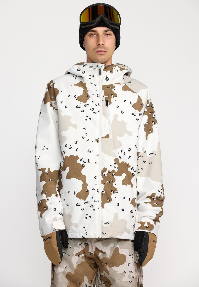 Camouflagejas in wit, bruin en beige met een ritssluiting aan de voorkant, verstelbare capuchon en elastische manchetten. Gecombineerd met bruine handschoenen.