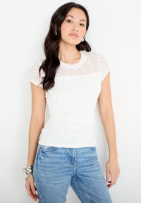 Joven mujer con el cabello largo y oscuro, vistiendo una blusa blanca de encaje y vaqueros azules, posando contra un fondo blanco liso.