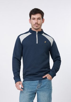 Férfi, aki sötétkék, negyedcipzáras pulóvert hord, szürke vállpanelekkel, valamint kék farmert, és egyszerű fehér háttér előtt áll.