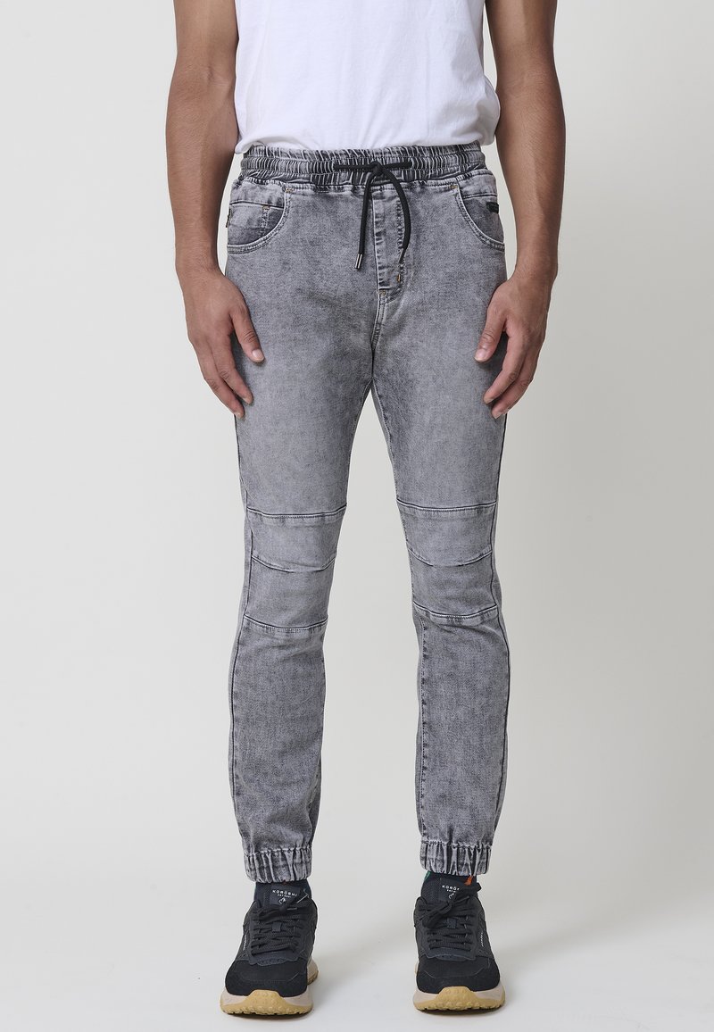 Pantalones jogger de mezclilla gris con cintura elástica, cierre de cordón y piernas ajustadas que presentan detalles en los paneles de las rodillas y puños.