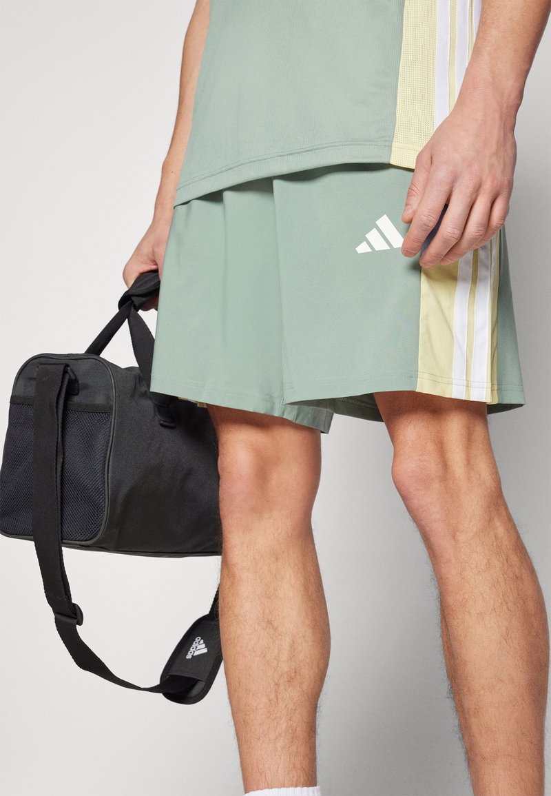 Pantalones cortos deportivos de color verde claro con acentos de tres franjas blancas, combinados con una bolsa deportiva negra. Tejido texturizado y diseño hasta la rodilla.